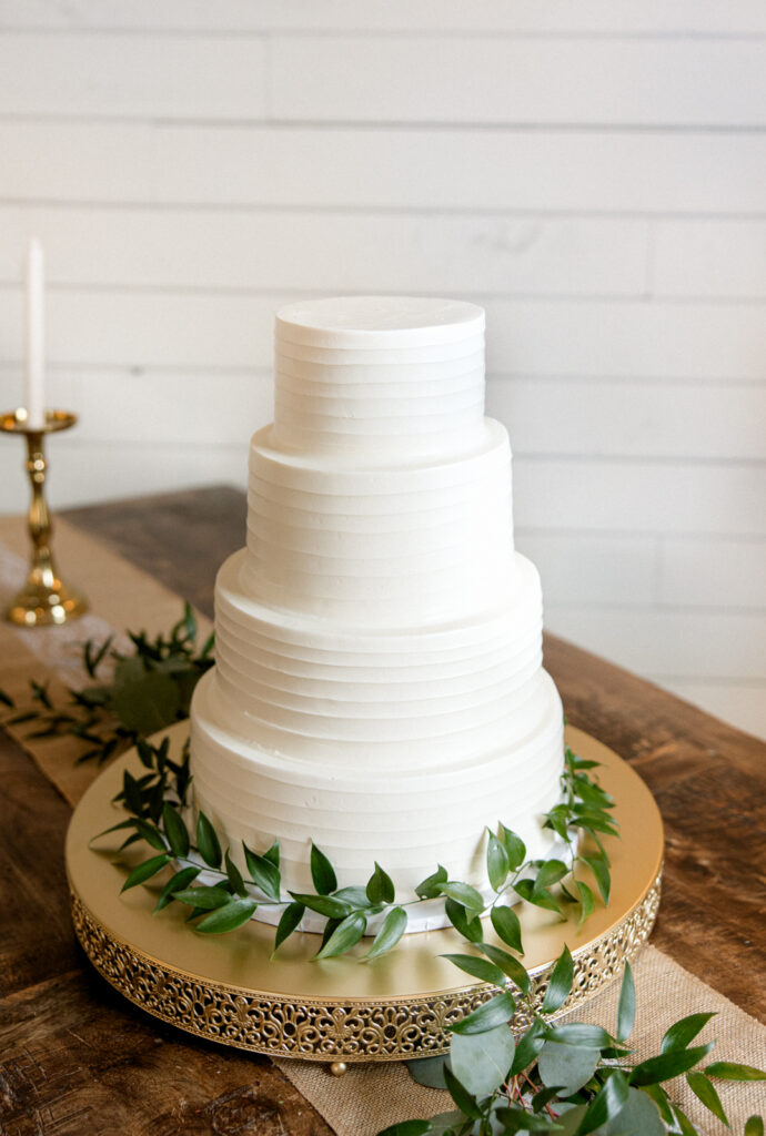Photo of classy white cake surrounded by greenery on a golden cake pedestal at Morgan Creek Barn in Dripping Springs, Texas. 