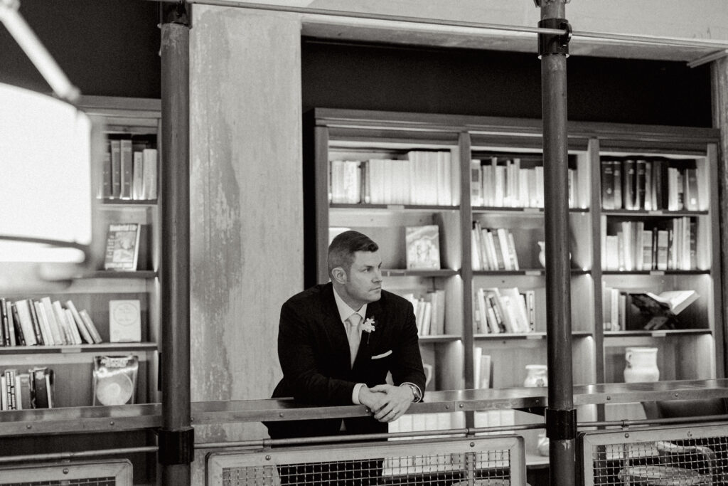 Groom leaning on railing with bookshelf behind him in Hotel Emma's Library.